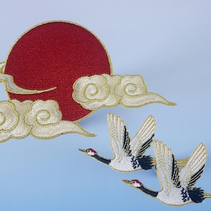 Op de afbeelding: Gestrikte strijkbare patches met een rode zon met witte wolken en twee witte kranen met zwarte accenten.