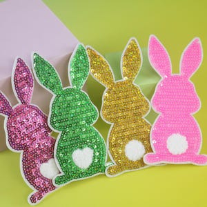 Könnte beinhalten: Vier paillettenbesetzte Hasenaufnäher in Rosa, Grün und Gold mit weißen Akzenten. Der grüne Hase hat ein weißes Herz. Die Aufnäher befinden sich auf einem gelben Hintergrund mit einer rosa Box.
