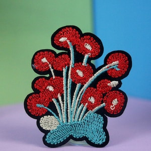 Può includere: Toppa ricamata con fiori rossi, contorno nero e base verde turchese. I fiori sono disposti a forma di bouquet.