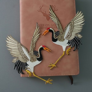 Peut inclure: Deux patchs brodés thermocollants représentant des grues blanches avec des accents noirs et dorés. Les grues sont en position de vol, les ailes déployées. Les patchs sont sur un carnet en cuir marron avec le texte "Keep on going, never give up".