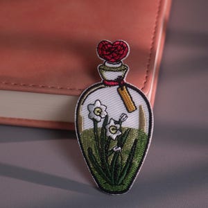 May include: Embroidered patch featuring a clear bottle design with white daffodils and green foliage. The bottle is topped with a red heart-shaped stopper and a small tag. The patch is set against a pink notebook.