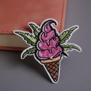 Könnte beinhalten: Gestickter Aufnäher mit einer rosa Eiswaffel mit grünen Cannabisblättern. Das Eis hat ein strukturiertes Aussehen mit tropfenden Details. Der Kegel ist braun mit einem Waffelmuster, und der Aufnäher hat einen weißen Rand.