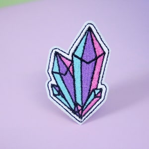 以下が含まれることがあります： 紫、ピンク、ブルーのクリスタルが刺繍された白いアイロン接着パッチです