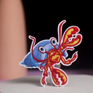 May include: Embroidered patch featuring a cartoon hermit crab. The crab has a blue shell, red claws, and yellow accents. The patch is white-bordered and appears to be made of fabric. The design is detailed and colorful, suitable for clothing or accessories.