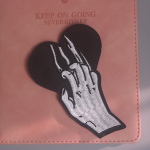 Peut inclure: Un écusson brodé en forme de cœur noir représentant une main squelettique blanche sur la surface. L'écusson est posé sur une couverture de carnet rose avec le texte en relief KEEP ON GOING NEVER GIVE UP visible en haut de la couverture.