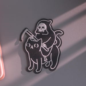 May include: A black and white embroidered patch featuring a grim reaper figure riding on the back of a black cat. The reaper holds a scythe, and the design is outlined in white thread against a black background, creating a high-contrast graphic.