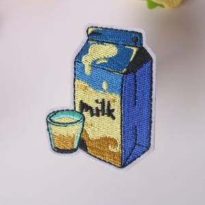以下が含まれることがあります： 青い牛乳パックと牛乳のコップが刺繍されたアイロン接着パッチ。牛乳パックには「Milk」と書いてあります。