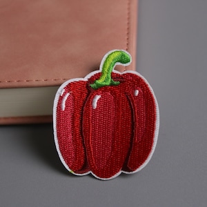 Può includere: Toppa ricamata raffigurante un peperone rosso con gambo verde e contorno bianco. La toppa è su una superficie grigia accanto a un taccuino rosa. Ideale per personalizzare abbigliamento o accessori.