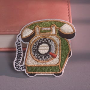May include: An embroidered patch featuring a vintage rotary telephone in shades of olive green, cream, and black. The design includes a coiled cord, a circular dial with black holes, and a small red indicator, all outlined with a white border.