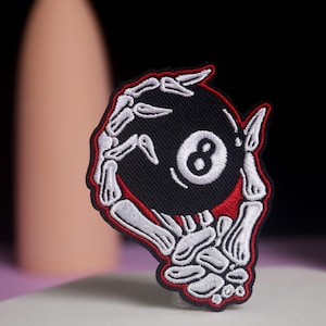 Könnte beinhalten: Gestickter Aufnäher mit einer schwarzen Achtkugel mit weißer Acht, gehalten von einer Skeletthand. Der Aufnäher hat einen roten Rand und weiße Nähte. Ideal für Jacken, Taschen oder andere Accessoires. Einzigartiges Design.