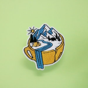 Puede incluir: Un parche bordado para planchar que presenta una taza amarilla con una escena de montaña en su interior. Las montañas son blancas con un río azul que fluye a través de ellas. Hay una pequeña tienda de campaña amarilla y un sol con rayos en la escena.