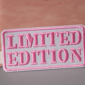 Könnte beinhalten: Ein rechteckiger Aufnäher mit den Worten "LIMITED EDITION" in rosa auf weißem Grund gestickt. Der Aufnäher hat einen rosa Rand und befindet sich vor einem verschwommenen rosa Notizbuch.