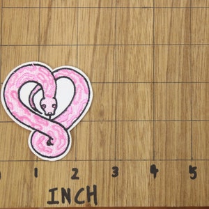Op de afbeelding: Roze geborduurde strijkpatch van een slang die in de vorm van een hart is opgerold. De patch is 5 cm breed en 5 cm hoog.