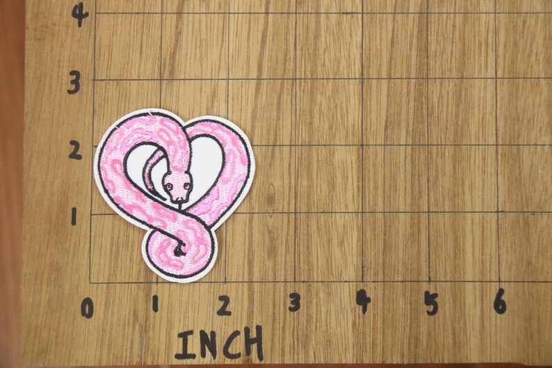Op de afbeelding: Roze geborduurde strijkpatch van een slang die in de vorm van een hart is opgerold. De patch is 5 cm breed en 5 cm hoog.