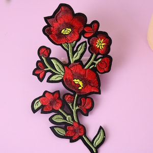 Könnte beinhalten: Rote gestickte Blumenapplikation mit grünen Blättern und schwarzen Umrandungen. Die Applikation ist perfekt, um Kleidung, Taschen oder anderen Accessoires einen Hauch von floralen Details zu verleihen.