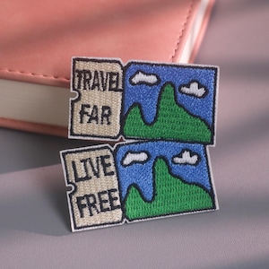 May include: Two rectangular embroidered patches featuring a landscape design. The top patch displays the text TRAVEL FAR, and the bottom patch displays the text LIVE FREE. Both patches show green mountains against a blue sky with white clouds.