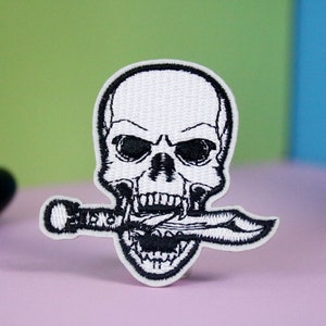 Puede incluir: Calavera bordada en blanco con un cuchillo negro en la boca. La calavera tiene una expresión amenazante con ojos negros y un contorno negro.
