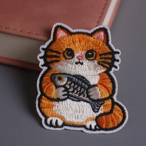 Op de afbeelding: Geborduurde patch met een cartoonkat met oranje en witte vacht, die een vis vasthoudt. De kat heeft grote ogen en een roze neus. De patch heeft een witte rand en is geschikt voor kleding of accessoires.