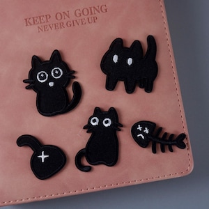 Può includere: Un quaderno rosa con il testo "Keep on going never give up" sulla copertina. Ci sono cinque toppe ricamate nere sulla copertina: un pesce, un gatto e tre gatti con occhi grandi.