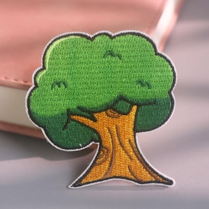 Puede incluir: Un parche bordado termoadhesivo con forma de árbol, con una copa de color verde vibrante y un tronco de color marrón dorado texturizado. El diseño presenta un borde negro grueso que perfila el follaje y el tronco, creando una representación gráfica clara de un árbol de hoja caduca.