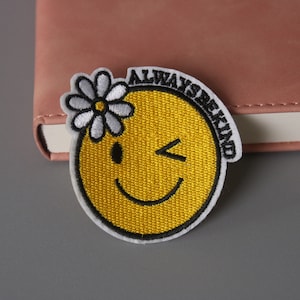 May include: Yellow embroidered iron-on patch with a winking smiley face and a white daisy on top. The text "Always Be Kind" is written around the smiley face.