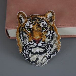 Könnte beinhalten: Nahaufnahme eines gestickten Tigergesichts-Patches. Der Patch ist braun, orange, schwarz und weiß. Der Tiger hat einen grimmigen Ausdruck mit grünen Augen.