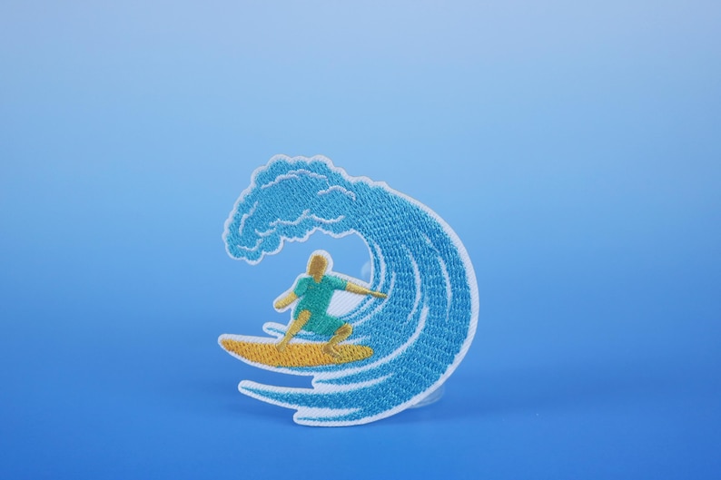 Może przedstawiać: Naszywka haftowana przedstawiająca surfer&oacute;w na fali. Fala jest niebieska i biała, a surfer ma na sobie niebieską koszulę i ż&oacute;łte spodenki. Naszywka znajduje się na niebieskim tle.