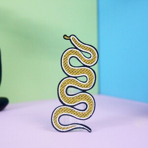 May include: A decorative, embroidered snake patch. The snake is yellow with a scale pattern and black outline. The patch is in a vertical, wavy design. The background is a mix of colors, including green, blue, and purple.