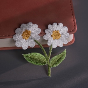 Puede incluir: Parche bordado de flores de margarita con pétalos blancos y centro amarillo, tallo y hojas verdes. El parche está colocado sobre un cuaderno de cuero marrón. El fondo es una superficie gris oscuro.