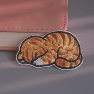 May include: An embroidered patch featuring a sleeping orange tabby cat with dark brown stripes, a white muzzle, and a white border. The cat is curled into a ball with its head tucked down, showcasing detailed thread work and a soft colour palette.