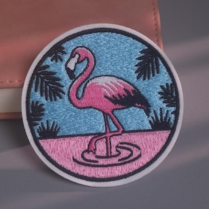 May include: A circular embroidered patch featuring a pink flamingo standing in pink water against a light blue background. The design includes dark blue palm leaf silhouettes and a thick white border, creating a detailed, colourful graphic emblem.