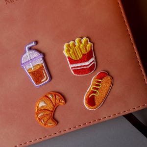 May include: Four embroidered iron-on patches featuring a croissant, a hot dog, french fries, and a cup of iced coffee. The patches are on a brown leather notebook.
