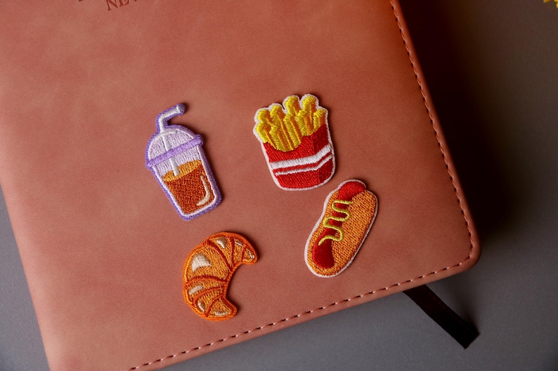 May include: Four embroidered iron-on patches featuring a croissant, a hot dog, french fries, and a cup of iced coffee. The patches are on a brown leather notebook.