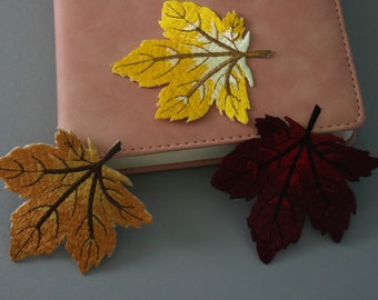 Può includere: Tre toppe di foglie in tessuto ricamate in colori giallo, arancione e bordeaux. Le toppe sono su un taccuino rosa.