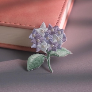 May include: An embroidered patch featuring a hydrangea flower with light purple and white petals, accompanied by two green leaves on a short stem. The embroidery uses dense thread work to create a textured floral design with clear color transitions.