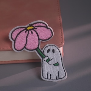 May include: An embroidered patch featuring a white ghost with black eyes holding a large pink flower with a yellow center and a green stem. The patch has a white border and is positioned against a pink notebook cover with a smooth texture.