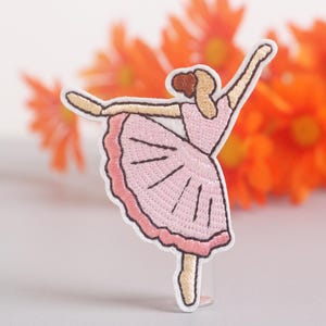 Puede incluir: Parche bordado de una bailarina con un vestido rosa, con los brazos extendidos. El parche tiene un borde blanco y se encuentra sobre un fondo borroso de flores naranjas. El pelo de la bailarina es castaño.