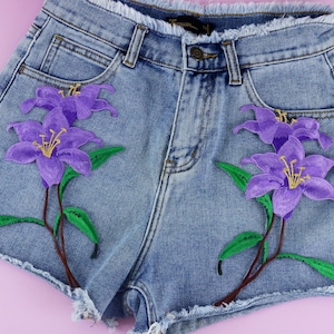 Könnte beinhalten: Hellblaue Jeans-Shorts mit lila bestickten Blumen. Die Blumen haben gelbe Zentren und grüne Stiele.
