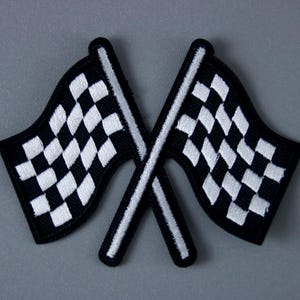 May include: Two embroidered racing flags with a black and white chequered pattern, crossed against each other. The flags have a black border and white flagpoles. The background is a solid grey.