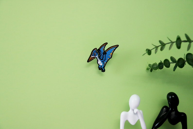 Pode incluir: Etiqueta bordada de ferro azul e amarela de um pterod&aacute;ctilo voando com as asas abertas.