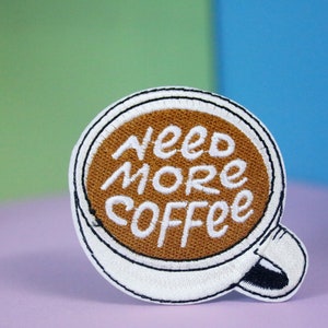 May include: White embroidered patch shaped like a coffee cup with brown coffee inside. The text "Need More Coffee" is embroidered in white on the brown coffee.