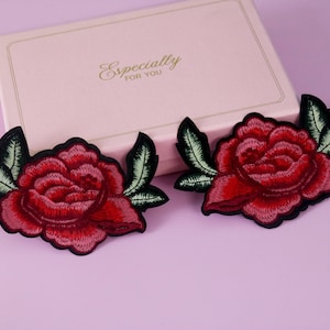 Peut inclure: Deux patchs brodés de roses rouges avec des feuilles vertes et des contours noirs. Les patchs sont sur une surface rose pâle.