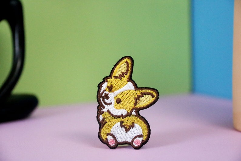 Może przedstawiać: Naszywka haftowana przedstawiająca brązowego i ż&oacute;łtego corgiego z białymi znaczeniami. Corgi patrzy w lewo i ma język wystający.