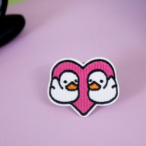 Könnte beinhalten: Gestickter Aufnäher mit zwei weißen Enten, die sich innerhalb eines rosa Herzens gegenüberstehen. Die Enten haben schwarze Augen und orangefarbene Schnäbel.