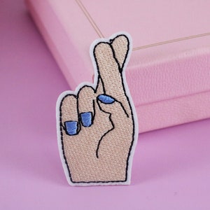 May include: A white embroidered patch of a hand with crossed fingers, with blue nail polish on the fingernails.