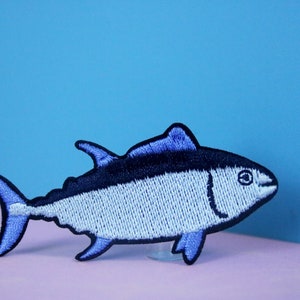 Puede incluir: Parche bordado con forma de pez, con tonos azules y negros. El pez tiene un diseño detallado con aletas, cola y ojo. El parche es probablemente para ropa o accesorios.