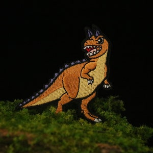 Puede incluir: Parche de dinosaurio bordado de color naranja y morado con una amplia sonrisa. El parche está sobre una superficie de musgo verde.