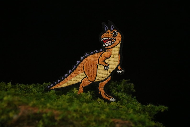 May include: Orange and purple embroidered dinosaur patch with a toothy grin. The patch is on a green mossy surface.