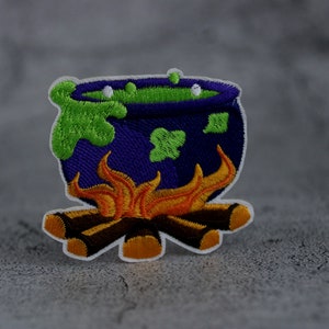 Puede incluir: Un caldero morado con líquido verde burbujeante se encuentra sobre un parche bordado blanco con llamas naranjas y troncos marrones.