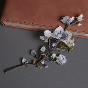 May include: An embroidered floral branch with grey and white flowers and green accents rests on a grey surface beside a brown leather-bound notebook. The delicate embroidery adds a touch of elegance.